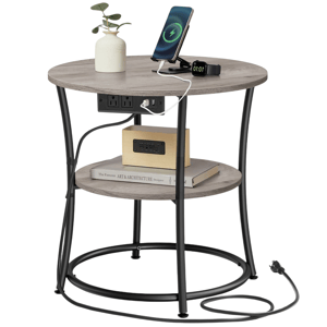 A round side table with black metal legs features a wooden top, two shelves, built-in power outlets and USB ports, and is shown with a charging phone and smartwatch, a small vase, books, and an alarm clock.