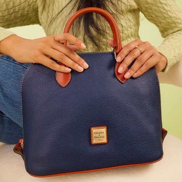 A navy blue pebbled leather handbag with brown handles and a gold logo plaque reading \“Dooney & Bourke.\“ A person wearing a green sweater and jeans holds the bag.