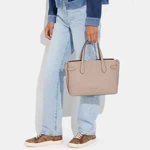 A person wearing blue jeans, a denim jacket, multi-colored sneakers, and holding a beige tote bag.