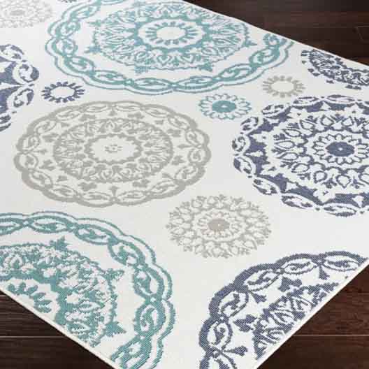 A Merriwood outdoor rug featuring circular, intricate patterns in teal, gray, and blue on a white background, placed on a dark wooden floor.