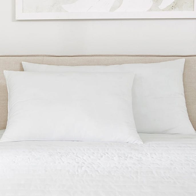 A bed features two large white pillows against a beige headboard, with white bedding and part of a decorative abstract print on the wall above.