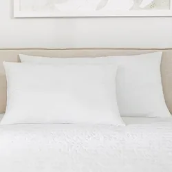 A bed features two large white pillows against a beige headboard, with white bedding and part of a decorative abstract print on the wall above.