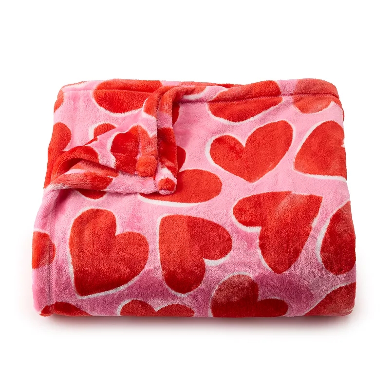 A pink oversized plush throw blanket features red heart patterns, offering a soft and cozy texture.