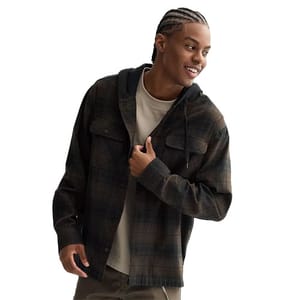 A man wears a dark plaid shacket with a hood, over a beige shirt, paired with olive pants. He has cornrow-style braids and is smiling, looking to the side.