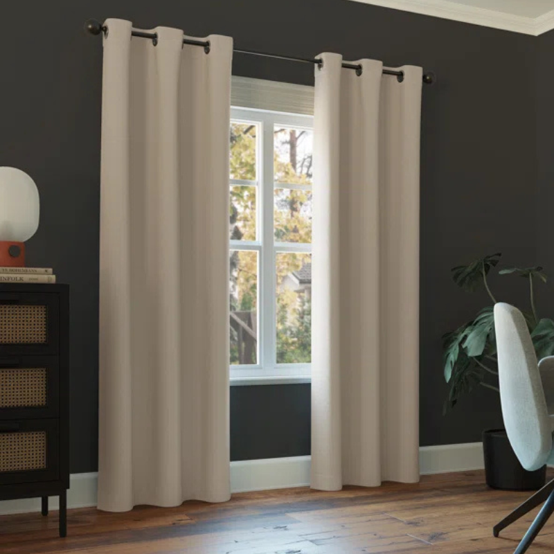 Beige blackout curtains hang from a rod in front of a window, set against a dark-colored wall. A chair and a plant are nearby, with light wooden flooring completing the room's decor.