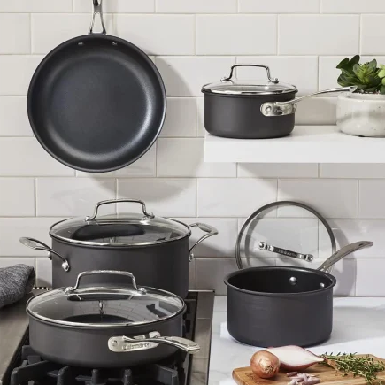 A set of dark nonstick cookware including a frying pan, saucepan, small pot, and large pot with lids, arranged on a kitchen counter and stove, with a cutting board nearby.