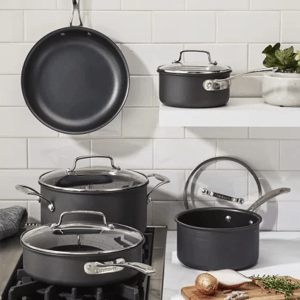 A set of dark nonstick cookware including a frying pan, saucepan, small pot, and large pot with lids, arranged on a kitchen counter and stove, with a cutting board nearby.