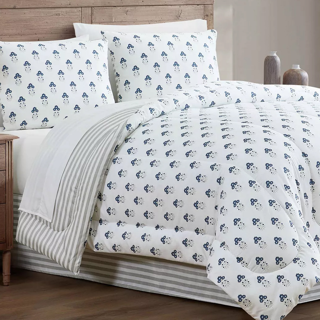 A bed features white bedding with a blue floral pattern, accompanied by matching pillows and a striped throw. The setting includes a wooden headboard and decorative vases on a nightstand.