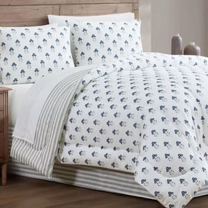 A bed features white bedding with a blue floral pattern, accompanied by matching pillows and a striped throw. The setting includes a wooden headboard and decorative vases on a nightstand.