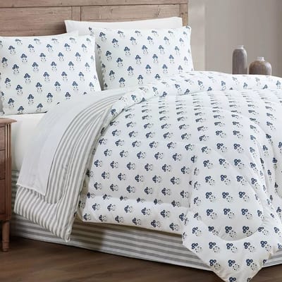 A bed features white bedding with a blue floral pattern, accompanied by matching pillows and a striped throw. The setting includes a wooden headboard and decorative vases on a nightstand.