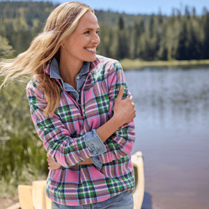 A woman wearing a plaid fleece jacket layered over a denim shirt, with a natural landscape and lake in the background.