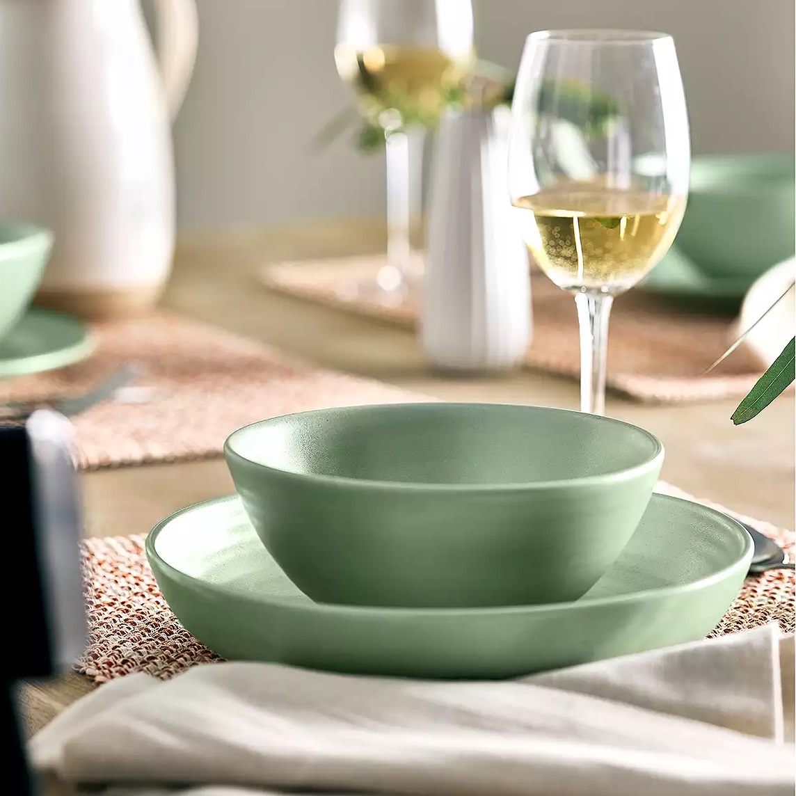 Green ceramic dinnerware set consisting of a plate, a deep plate, and a bowl on a table with a glass of white wine.