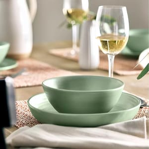 Green ceramic dinnerware set consisting of a plate, a deep plate, and a bowl on a table with a glass of white wine.