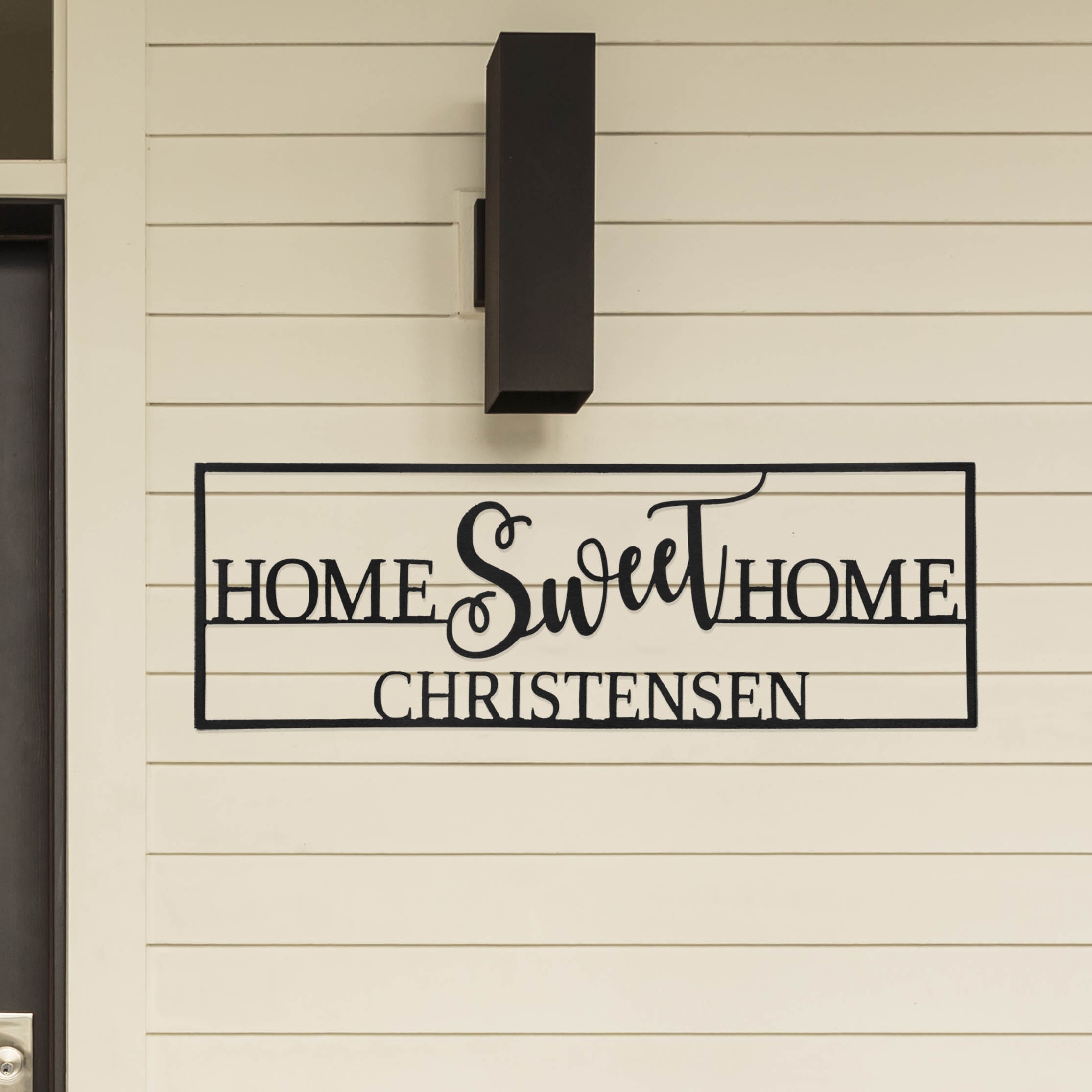 A black metal sign on a beige wall reads \“Home Sweet Home Christensen\“ featuring a decorative script for the word \“Sweet,\“ positioned under a modern black outdoor light fixture.