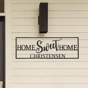 A black metal sign on a beige wall reads \“Home Sweet Home Christensen\“ featuring a decorative script for the word \“Sweet,\“ positioned under a modern black outdoor light fixture.