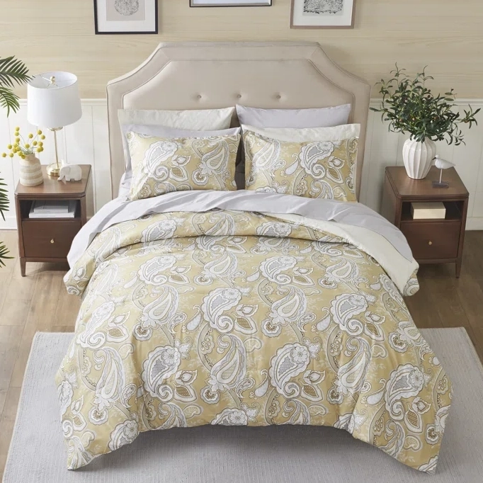 A paisley patterned bedding set with comforter, pillows, and sheets in gold, white, and gray tones arranged on a bed.