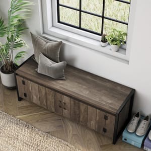The Oak 49\“ Iron Legs Storage Bench features a rustic wooden design with dual doors and black metal legs, offering storage and a surface for seating. Two decorative pillows are placed on top, and shoes are stored underneath.