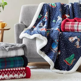 Holiday Lane Cozy Plush Throws feature festive patterns including plaid, snowflakes, and Christmas trees, with a warm sherpa lining. They are draped over a chair and stacked nearby, offering a cozy, seasonal look.