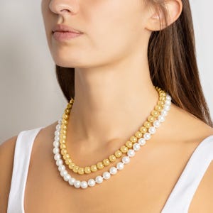 A woman is wearing two layered necklaces, one with gold beads and the other with white pearls. She is dressed in a white tank top.