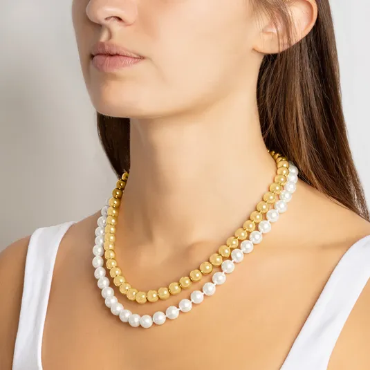 A woman is wearing two layered necklaces, one with gold beads and the other with white pearls. She is dressed in a white tank top.
