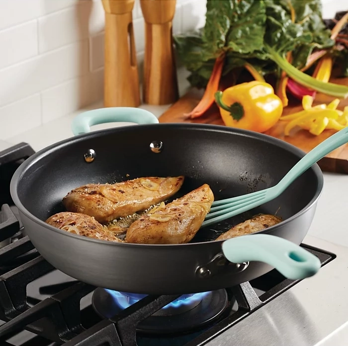 Non-stick frying pan with blue handles cooking chicken breasts on a gas stove, set against a backdrop of kitchen utensils and colorful vegetables.