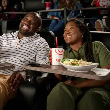 A couple enjoys a meal and drink in a movie theater with reclining seats and attached trays, featuring branded cups and plates.