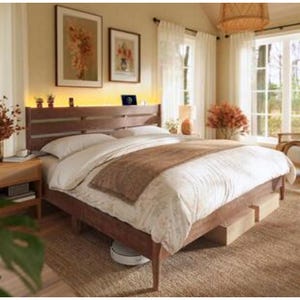 A cozy bedroom features a wooden bed frame with a white and beige bedding set, a Roomba under the bed, a bedside table with decor, framed wall art, and natural light streaming through white curtains.