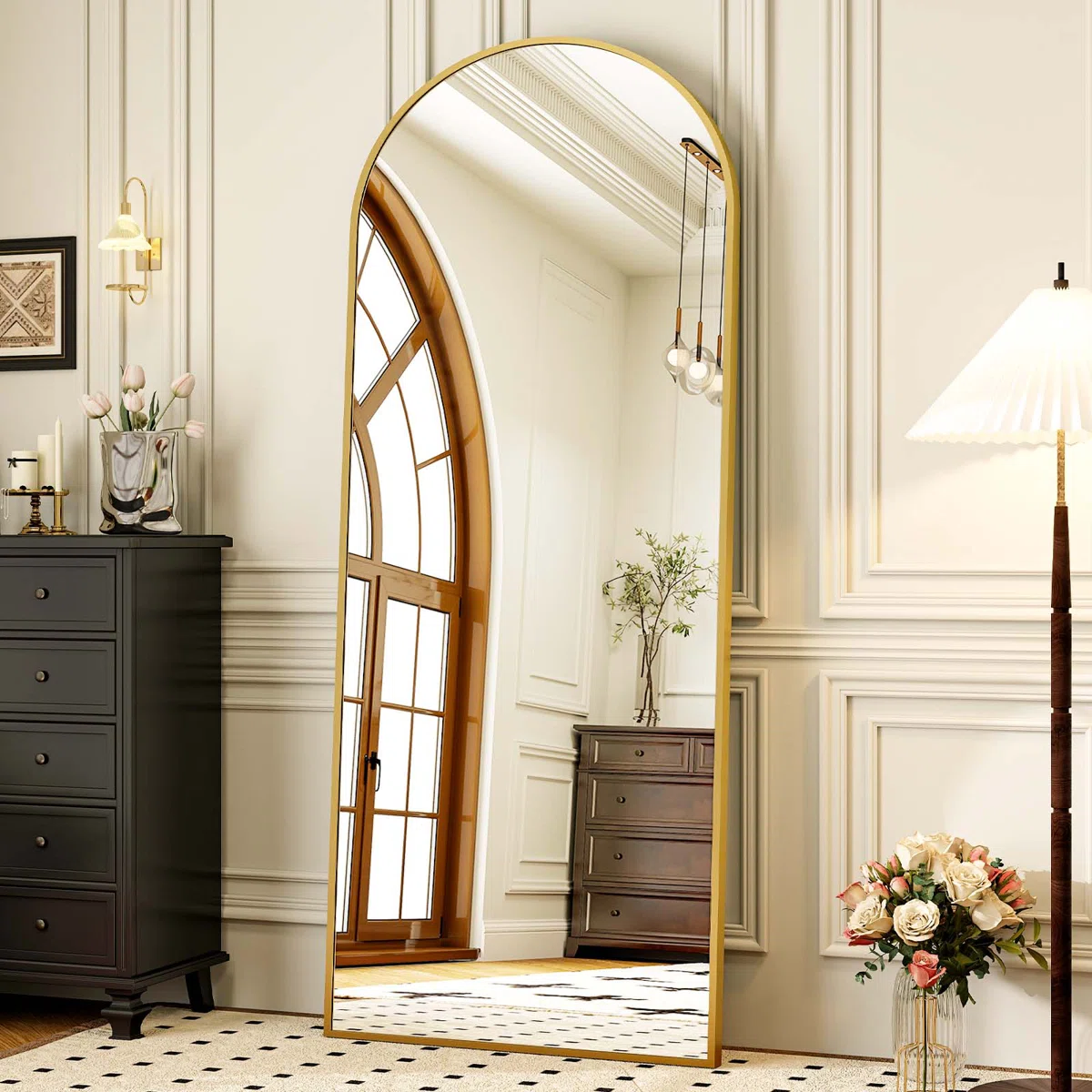 A tall, arched mirror with a gold frame reflects an elegant room with a window, black chest of drawers, and a vase with flowers. A floor lamp and vase of flowers are next to the mirror on patterned carpeting.