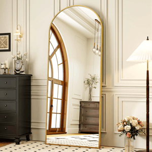 A tall, arched mirror with a gold frame reflects an elegant room with a window, black chest of drawers, and a vase with flowers. A floor lamp and vase of flowers are next to the mirror on patterned carpeting.