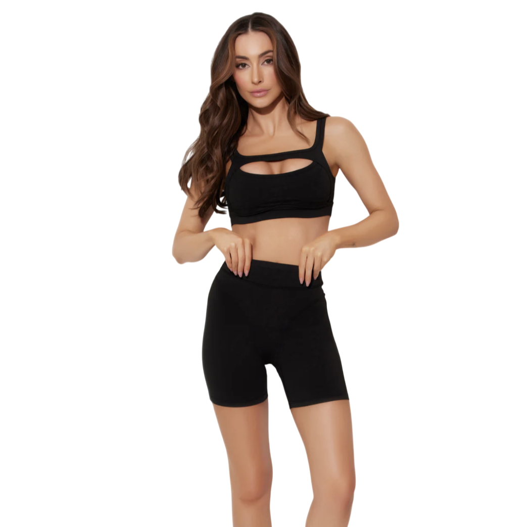 A woman in a black two-piece activewear set, featuring a sports bra with a cutout design and high-waisted shorts, paired with black athletic shoes, stands against a plain background.