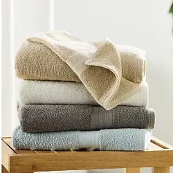 A stack of four neatly folded towels in beige, white, gray, and light blue colors is placed on a wooden stool, against a softly lit background with some greenery.
