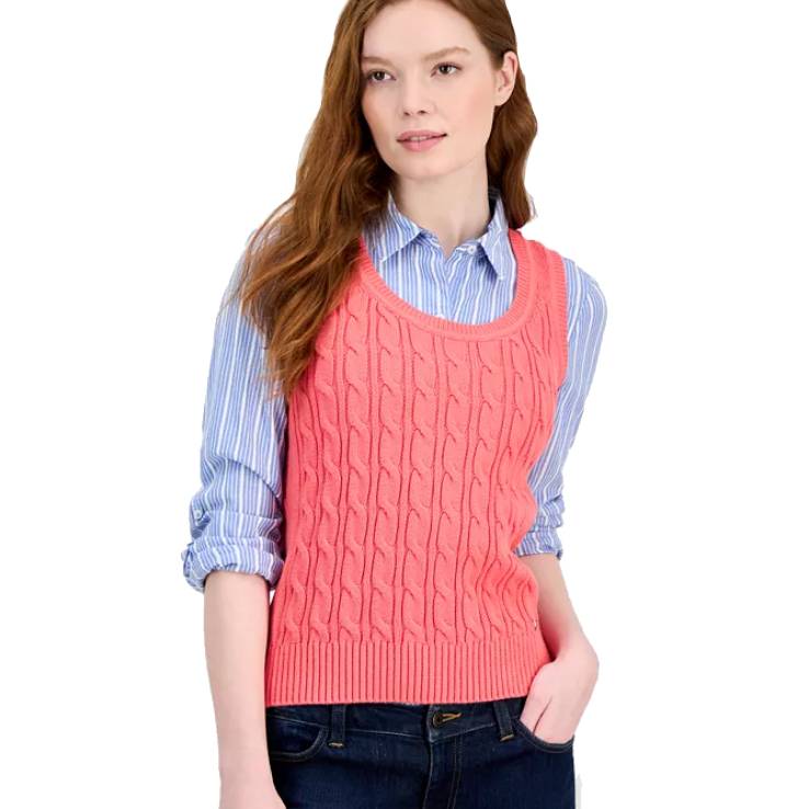 A woman is wearing a pink cable knit vest over a blue and white striped button-up shirt with rolled sleeves. She pairs them with dark jeans.