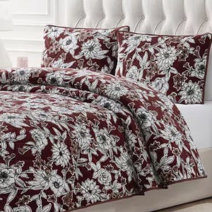 A bed is dressed with a maroon floral-patterned quilt and two matching pillow shams. The design features intricate white flowers on a dark background.