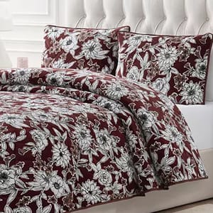 A bed is dressed with a maroon floral-patterned quilt and two matching pillow shams. The design features intricate white flowers on a dark background.