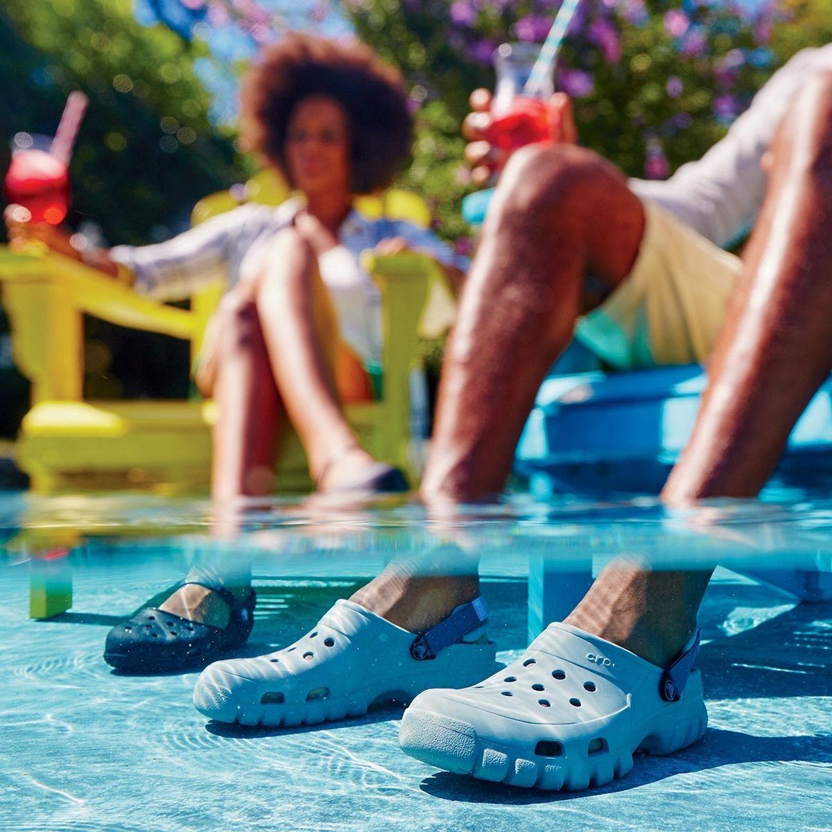 A pair of blue and a pair of gray foam clog shoes by a poolside, with a blurred background of people lounging.