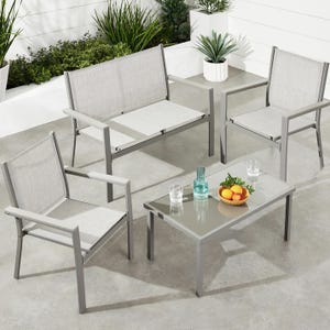 Outdoor patio set with two single chairs, a loveseat, and a glass-top coffee table. The furniture features a minimalist design with a grey metal frame and light grey fabric. The table holds a glass bottle, glasses, and a bowl of fruit.