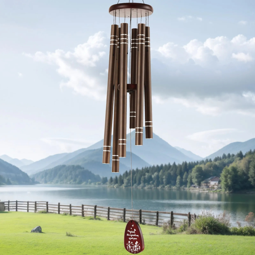 Bronze-colored wind chimes with a wooden top hang in a scenic outdoor setting by a lake, with mountains in the background.