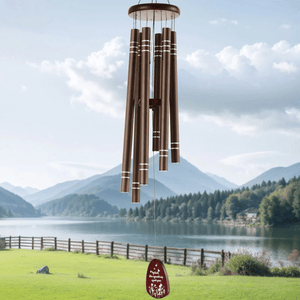 Bronze-colored wind chimes with a wooden top hang in a scenic outdoor setting by a lake, with mountains in the background.