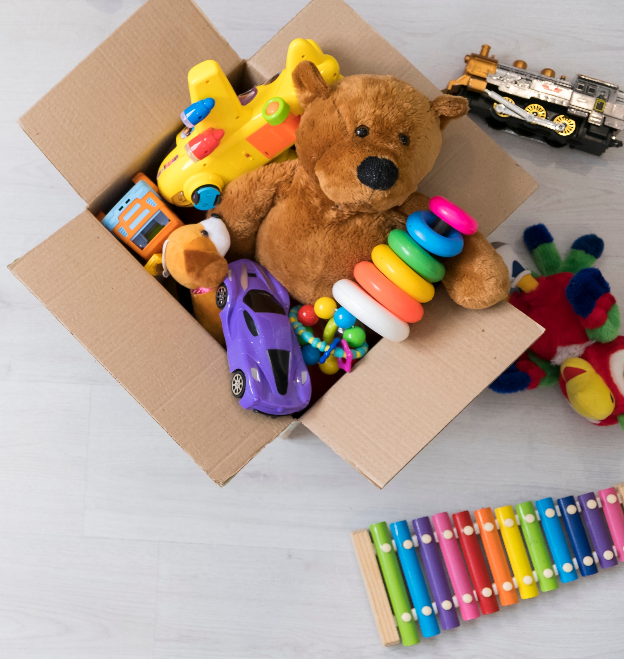 A cardboard box with assorted children’s toys including a stuffed bear, toy cars, a colorful xylophone, and a plastic ring stacker.