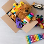 A cardboard box with assorted children’s toys including a stuffed bear, toy cars, a colorful xylophone, and a plastic ring stacker.