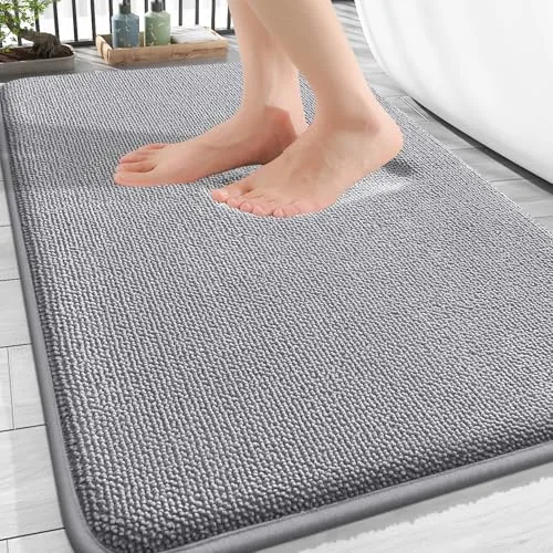 A person's feet are stepping onto a gray textured bath mat placed on a tiled floor next to a bathtub.