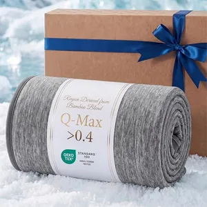A gray bamboo blend blanket labeled \“Q-Max >0.4\“ with OEKO-TEX certification is rolled up on a snowy surface next to a brown gift box tied with a blue ribbon.
