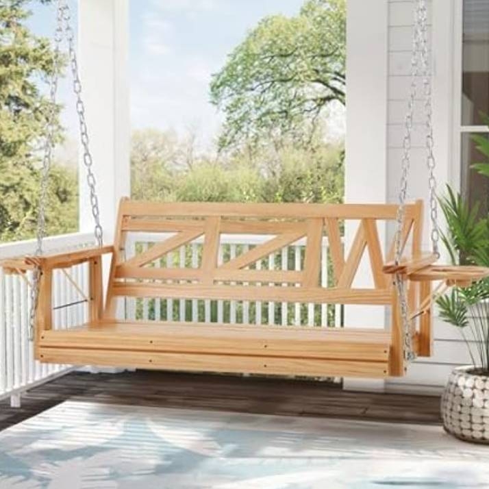 A wooden porch swing with geometric backrest design hangs with metal chains from a ceiling on a covered porch. The swing is surrounded by a railing with green foliage and has armrests on both sides.