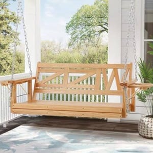 A wooden porch swing with geometric backrest design hangs with metal chains from a ceiling on a covered porch. The swing is surrounded by a railing with green foliage and has armrests on both sides.