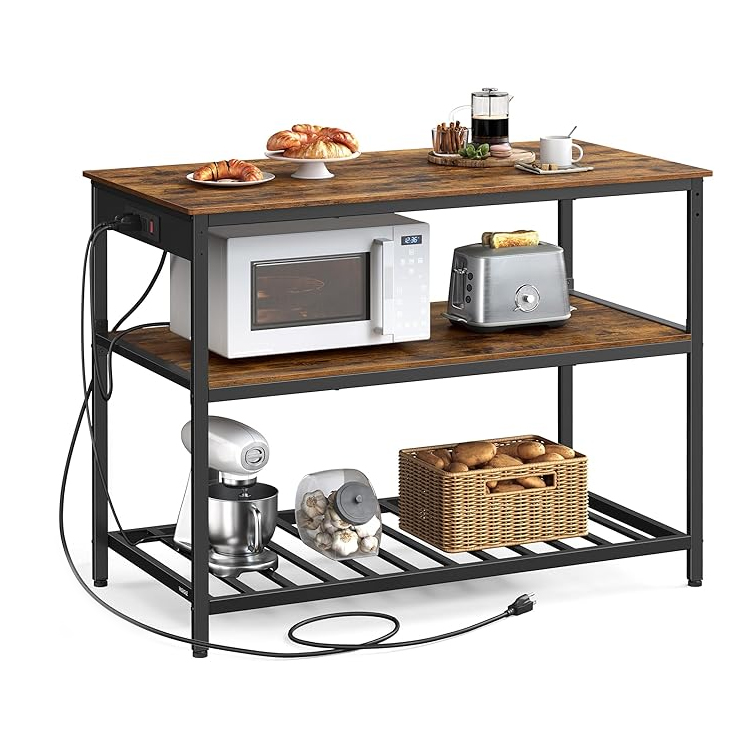 Kitchen island with three shelves; top holds croissants, a coffee maker, and a cup, middle has a microwave and toaster, bottom contains a stand mixer, a jar of garlic, and a basket of potatoes. Black metal frame, wooden surfaces, and built-in power strip.