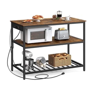 Kitchen island with three shelves; top holds croissants, a coffee maker, and a cup, middle has a microwave and toaster, bottom contains a stand mixer, a jar of garlic, and a basket of potatoes. Black metal frame, wooden surfaces, and built-in power strip.