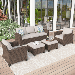 Outdoor patio furniture set with a wicker sofa, two armchairs, two ottomans, and a small table, all with gray cushions, set on a patterned outdoor rug, surrounded by greenery.