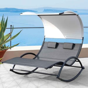 A double lounge chair with a curved frame and canopy, featuring two headrests, set on a terrace overlooking a body of water, accompanied by a potted plant.