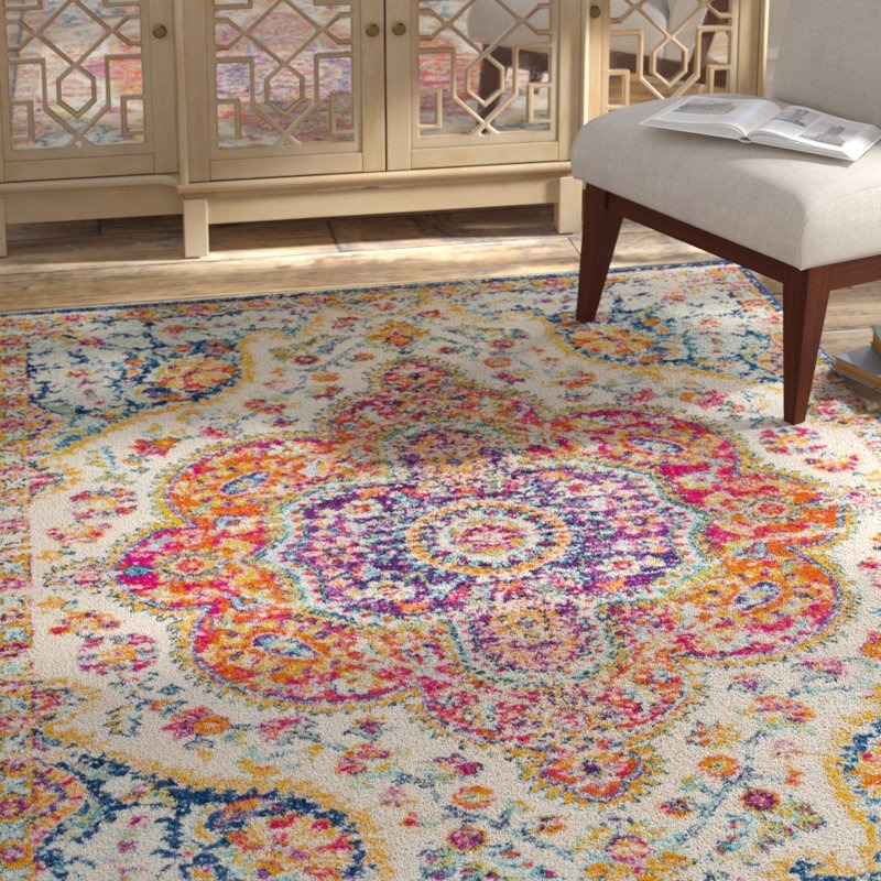 A colorful, ornate area rug with intricate patterns in pink, orange, yellow, and blue sits on a wooden floor. A light gray chair with wooden legs is nearby, accompanied by an open book.