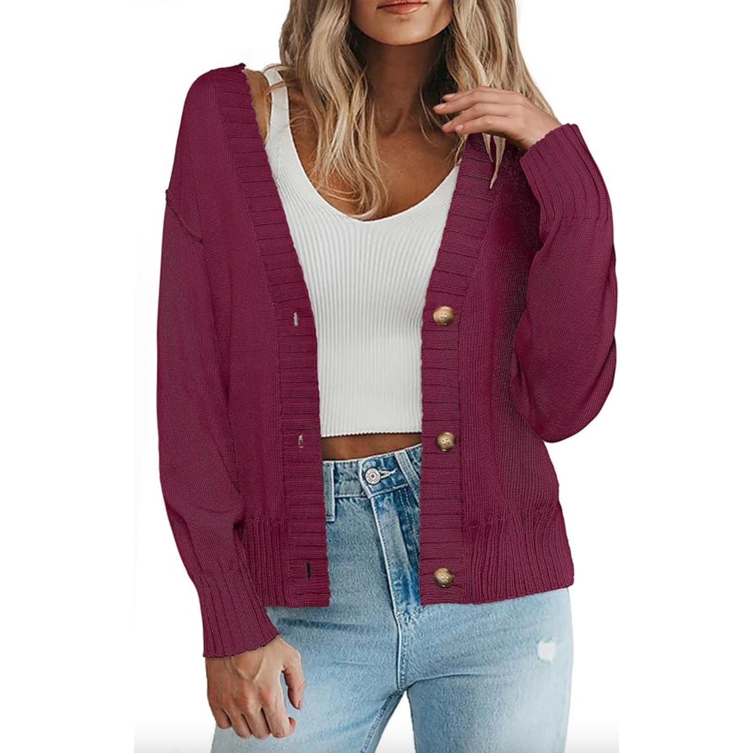 A woman is wearing a burgundy cardigan with button details over a white ribbed tank top and light blue jeans.